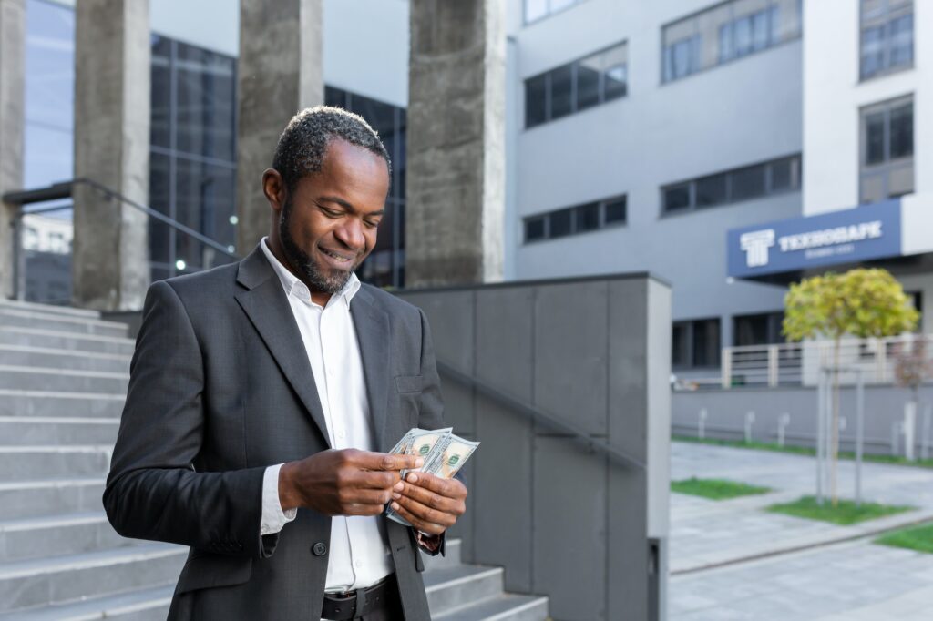 Happy African-American man, office worker stands near office, holds cash in hands, counts money